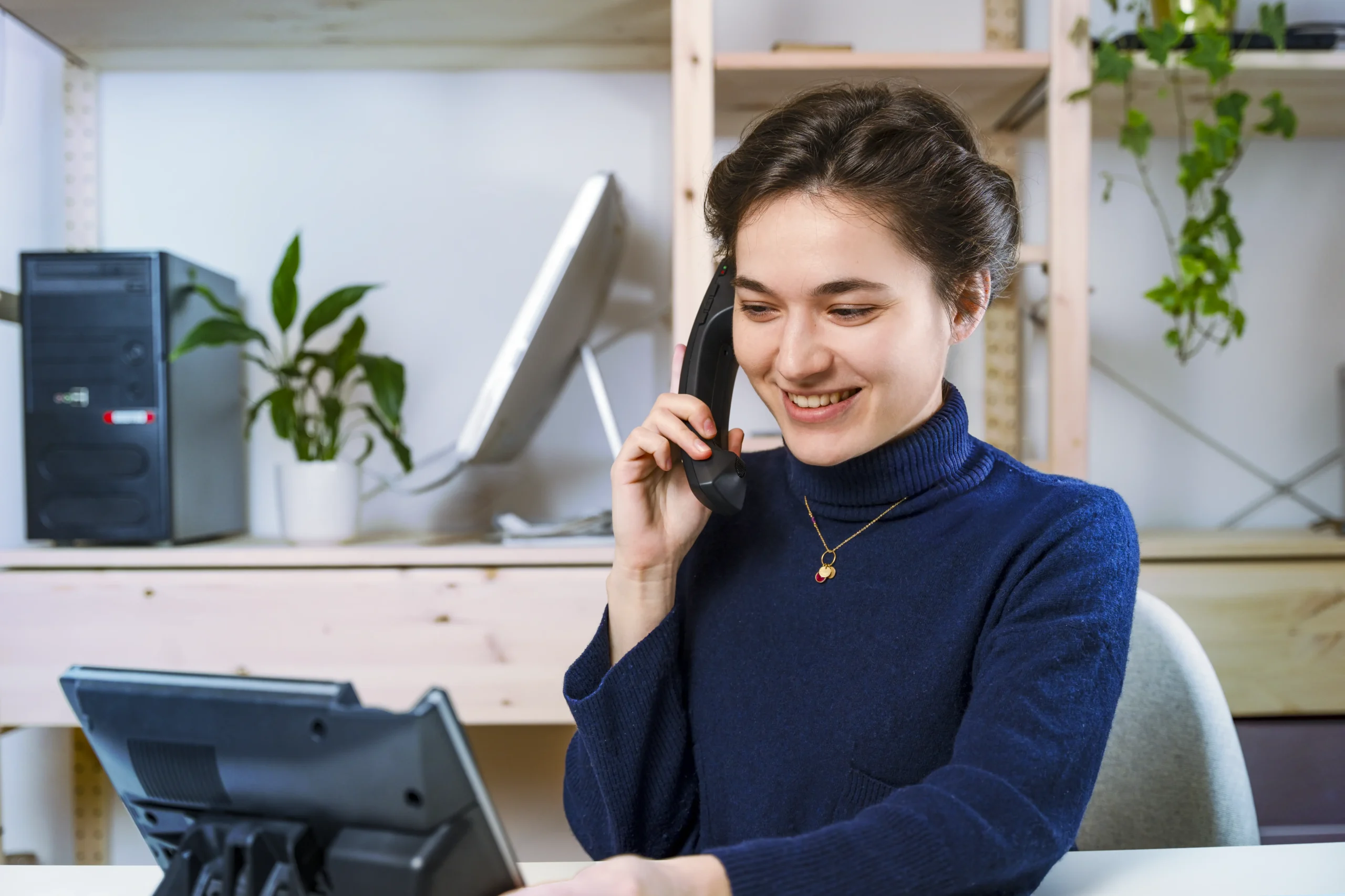 friendly IT secretary in a blue collar jumper on the phone, surrounded by computers and plants on the shelf