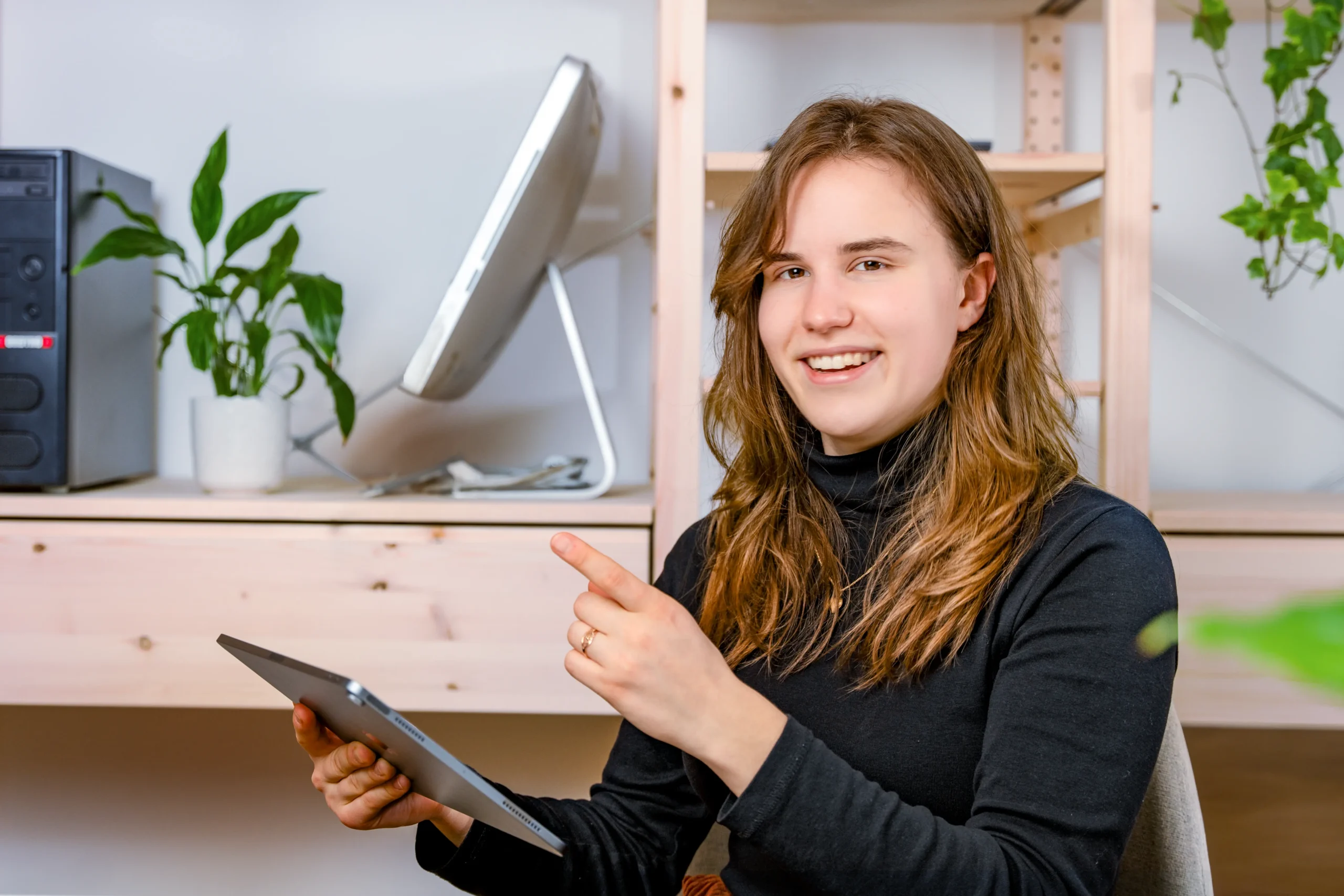 une aimable employée de IT-Dokter avec un Ipad pointe son index vers la gauche, entourée d'ordinateurs et de plantes sur l'étagère