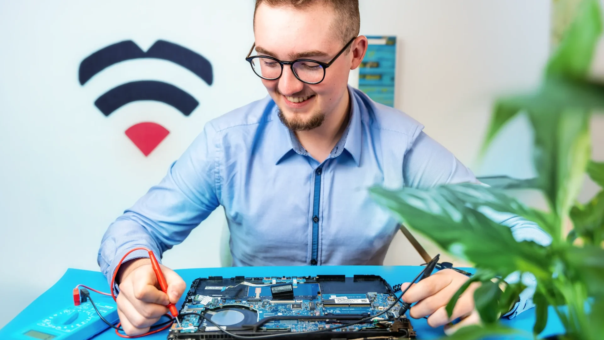 smiling IT-Dokter expert in blue shirt, carrying out a laptop repair, plant in the background, visible IT-Dokter logo